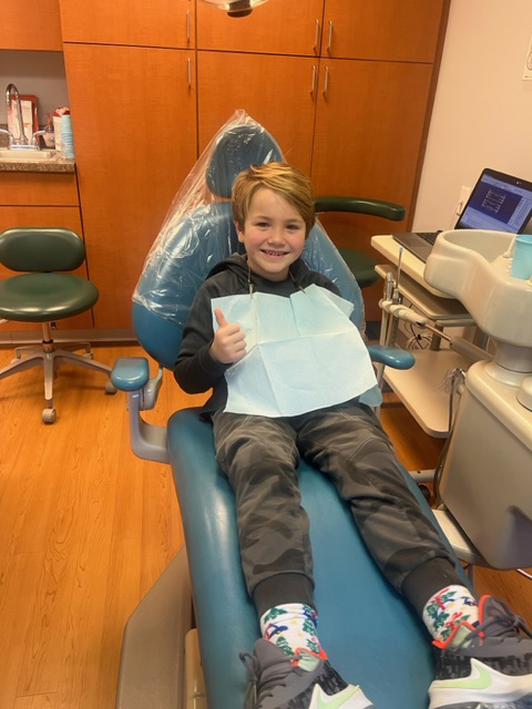 A young boy sitting in a dental chair with a smile on his face, wearing a blue mask and gloves, surrounded by dental equipment.