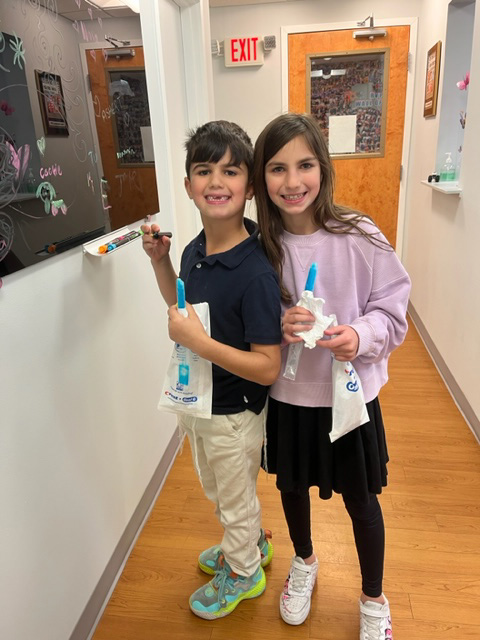 The image shows two individuals, likely children, standing together in an indoor setting with a bulletin board and chalkboard in the background. They are holding toothbrushes and appear to be participating in an activity related to dental hygiene.