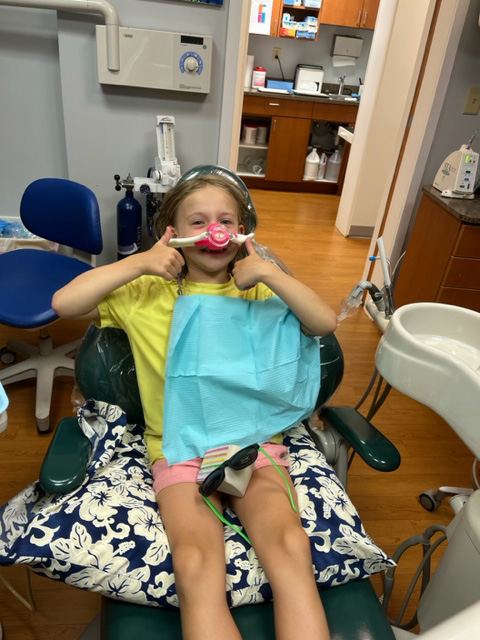 A young girl sitting in a dental chair with a mask over her mouth and nose, posing for the camera with a thumbs-up gesture.