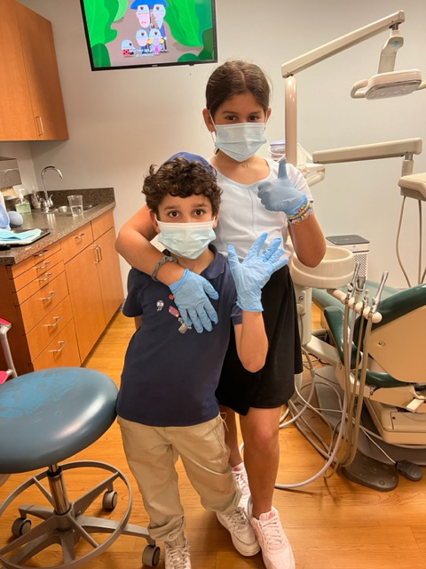 In this image, two individuals are standing in front of dental equipment, with one person wearing a face mask and gloves, posing for the photo.