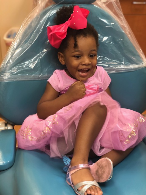 A small child sitting in a dental chair, smiling at the camera, with a pink tutu and a bow on their hair.