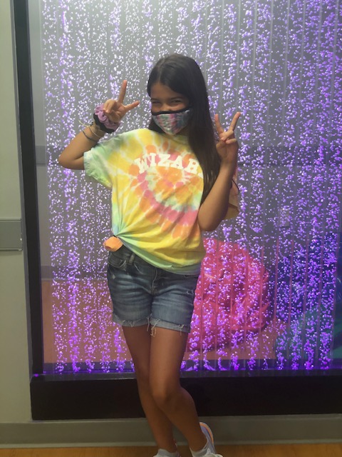 The image shows a person standing in front of a colorful wall with a peace sign on their hand, smiling at the camera. They are wearing a tie-dye t-shirt, shorts, and sneakers.