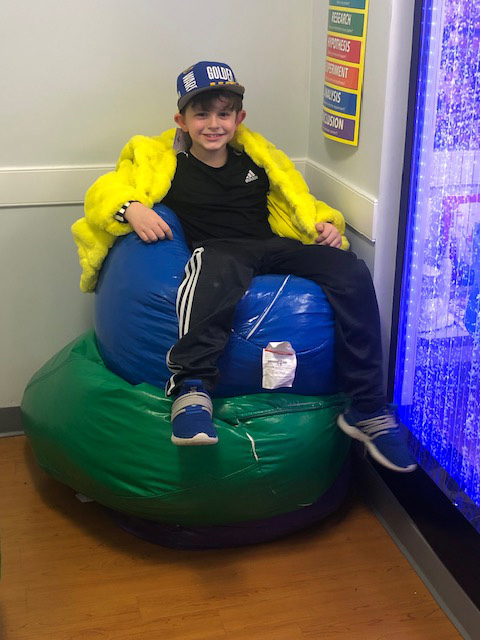 A young boy sitting on a large blue bean bag chair with a yellow jacket draped over him, wearing a cap, in an indoor setting.