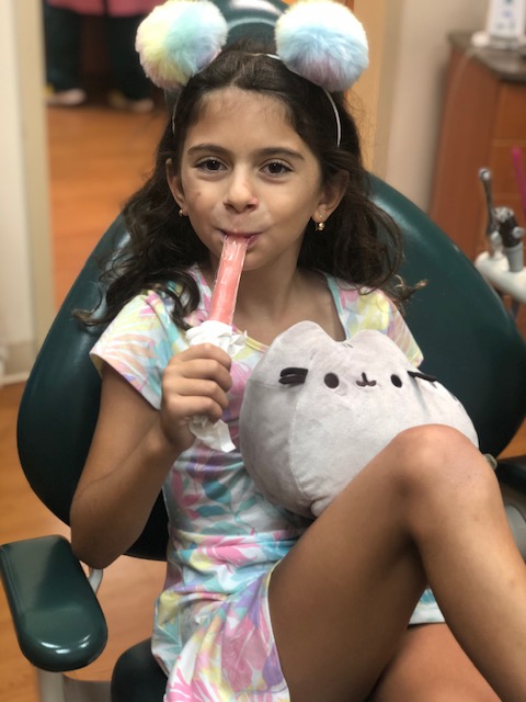 A young girl with a stuffed animal, sitting in a dental chair, eating a popsicle while looking at the camera.