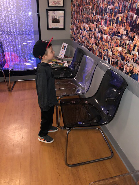 The image shows a young boy standing next to a wall covered with photographs, looking at them while wearing a baseball cap.