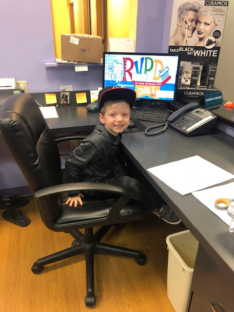 A young boy sitting at a desk with a computer monitor behind him, wearing a baseball cap, in an office setting.