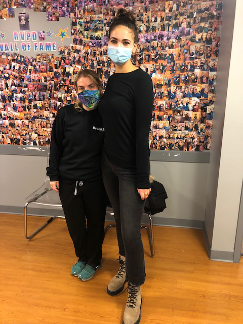 This is a photograph featuring two individuals standing indoors; one person appears to be wearing a black t-shirt, a face mask, and has their hair pulled back into a bun, while the other individual is dressed in a black top with a white logo on the chest, dark pants, and a face mask. They are positioned near a wall adorned with photographs and text, suggesting an event or exhibition setting. The environment includes a collage of images, possibly related to the theme of the gathering, but it's not clear from this image alone what the specific content of these photos is.