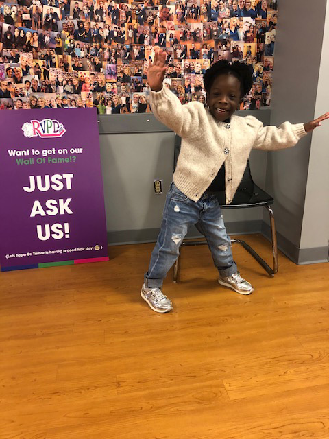 A young child with a raised hand is standing next to a purple sign with white text on a wall adorned with photos, smiling towards the camera.