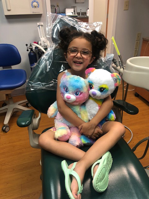 Girl sitting on dental chair holding two teddy bears.
