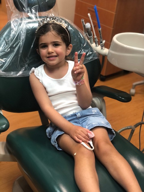 A young girl sitting in a dental chair, smiling and giving a peace sign with her right hand.