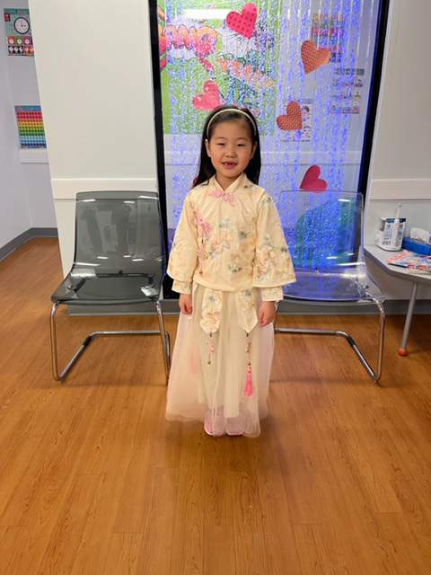 A young girl wearing traditional Asian clothing stands in front of a colorful display with a heart-shaped cutout.