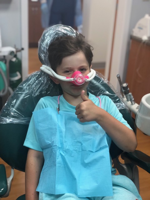 A young boy wearing a medical mask and sitting in a dental chair, giving a thumbs up sign with his right hand.