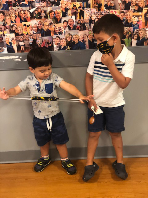 Two young boys standing next to each other, with one boy wearing a face mask and holding a string attached to a balloon, while the other boy has his back turned towards the camera.