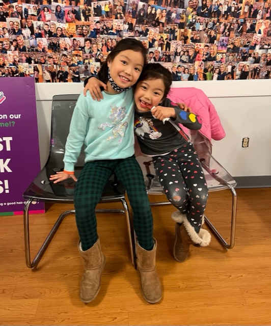 The image shows two young girls posing for a photo together, with one girl sitting on a chair while the other stands behind her. They are indoors, likely at an event, indicated by a sign in the background that reads 'Welcome our new team members.