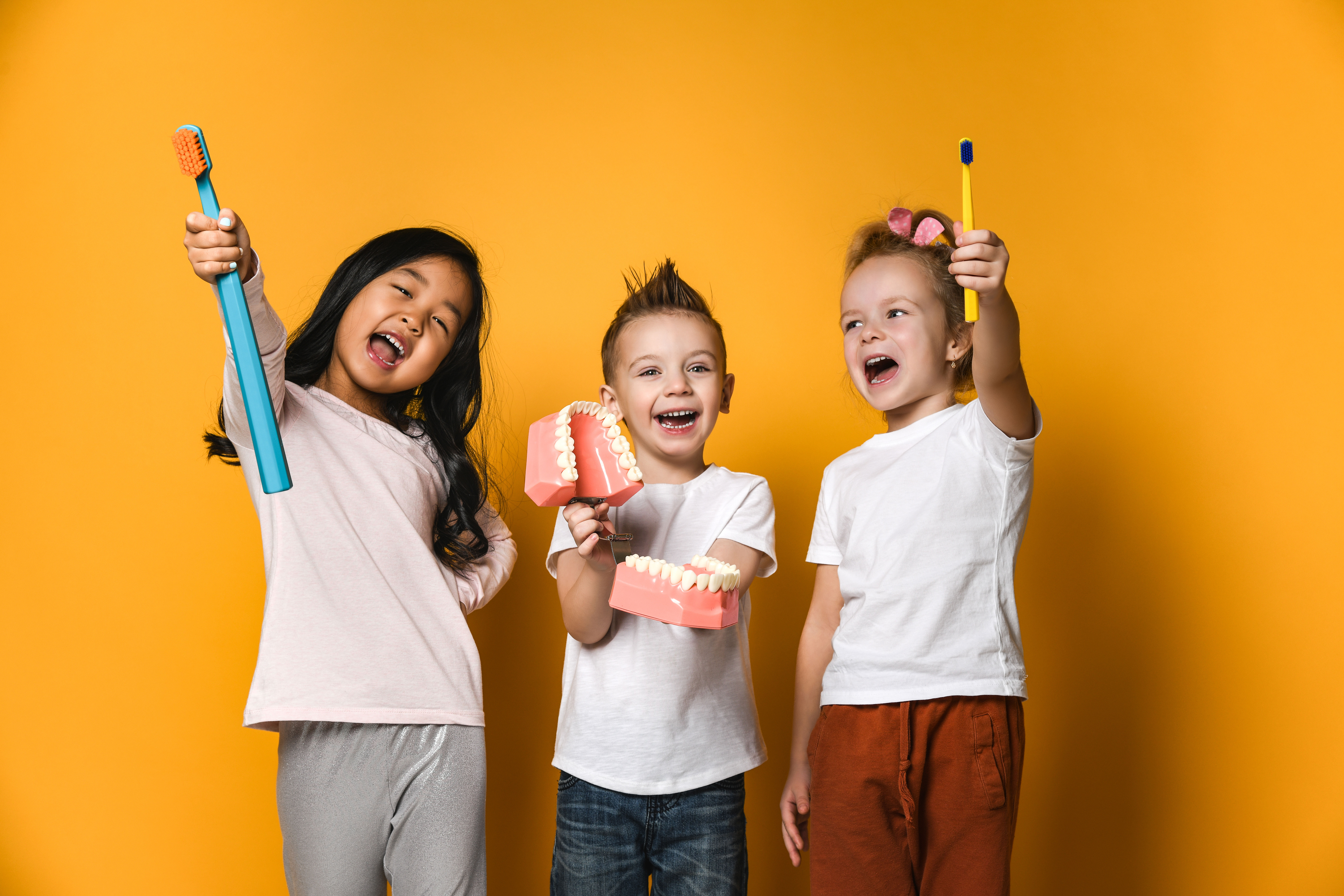 Three children holding toothbrushes in front of yellow background.