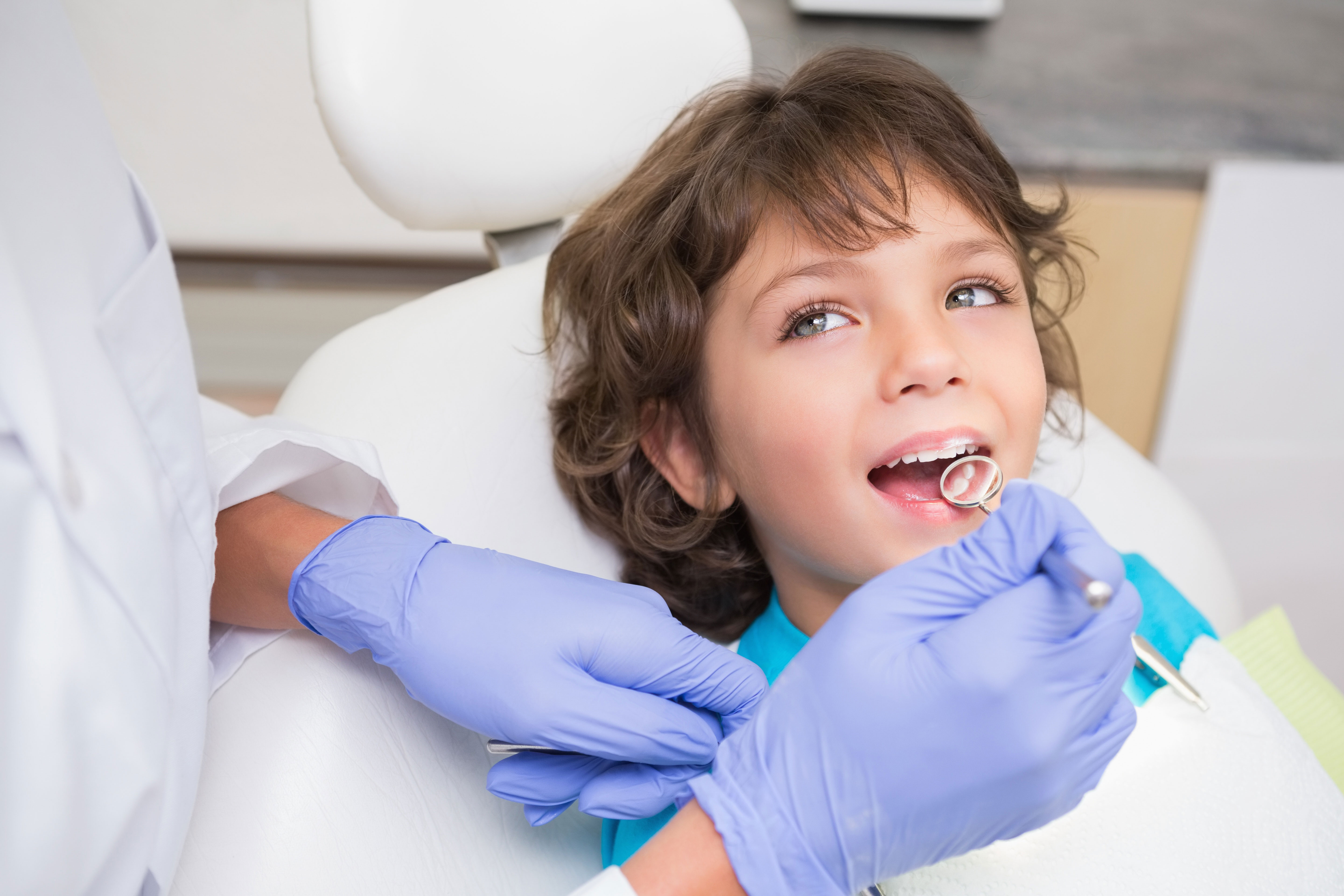 The image shows a young child sitting in a dental chair, smiling with their mouth open while receiving dental care from an adult dentist wearing protective gloves.