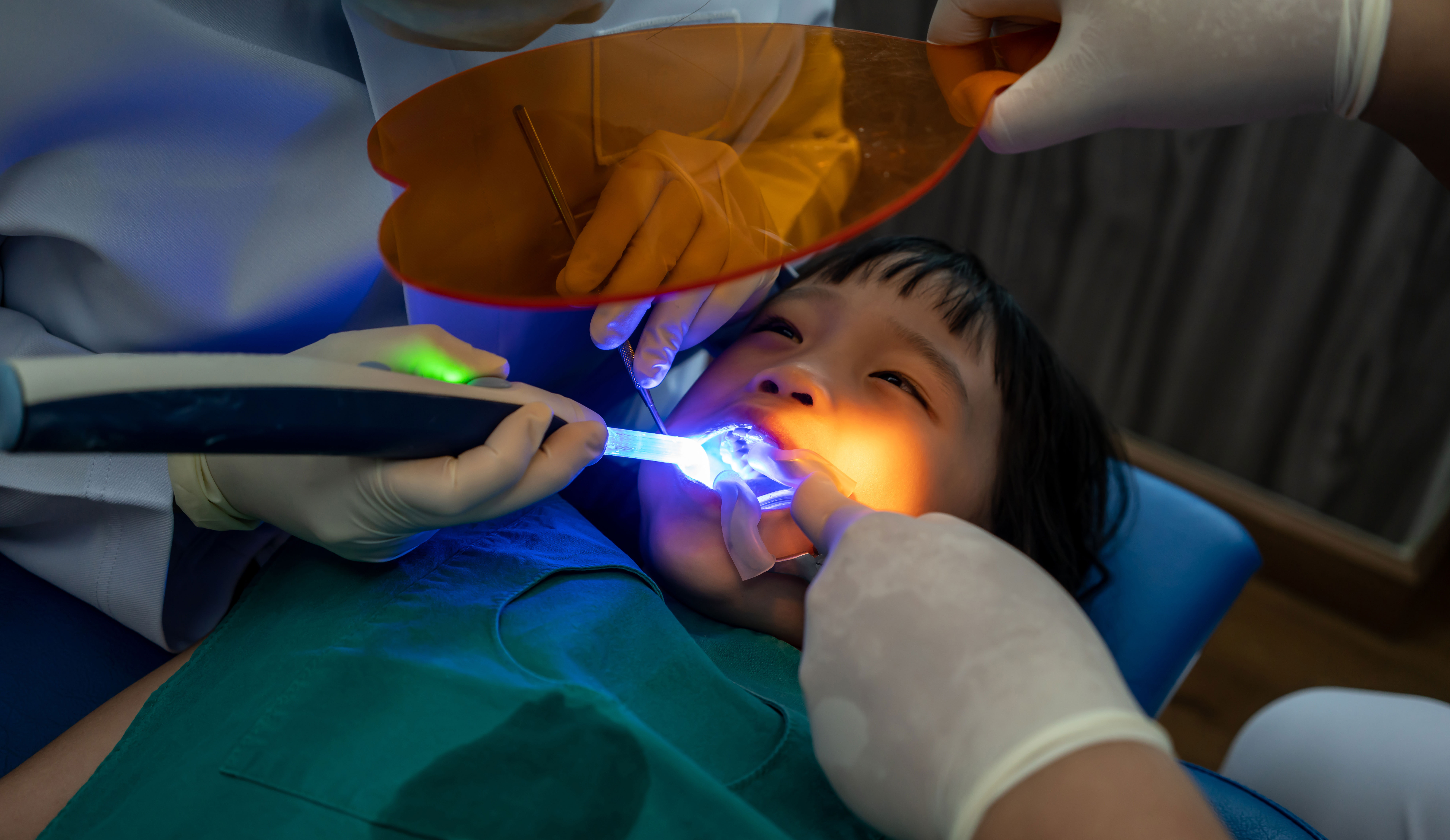 The image shows a dental care scene with a person receiving treatment while wearing a blue surgical gown, with a dental professional using a dental drill on their teeth, and a green laser device being used by another dental professional.