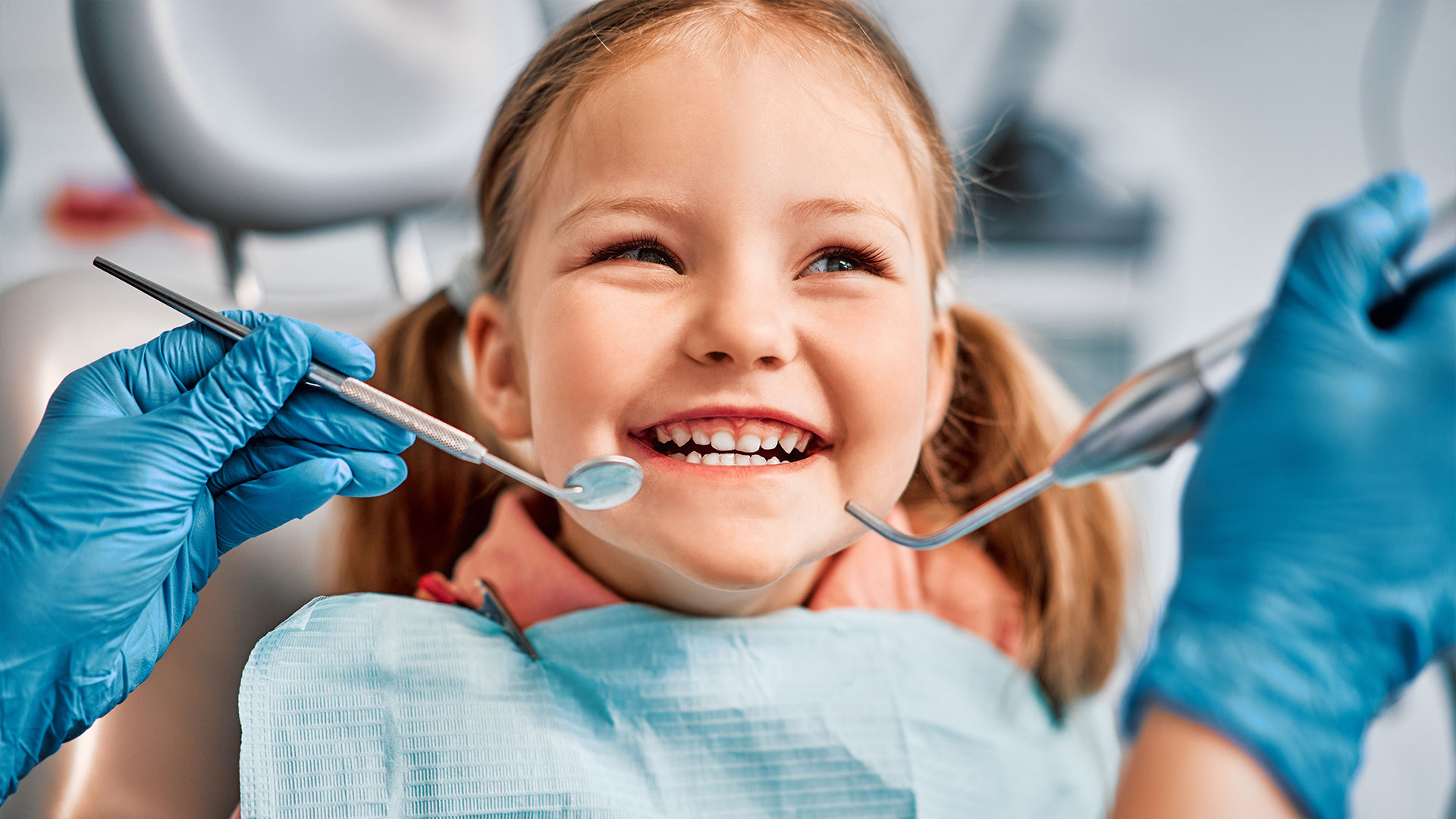A young girl with a broad smile is seated at a dental chair, wearing blue gloves and holding dental tools, while looking towards the camera.
