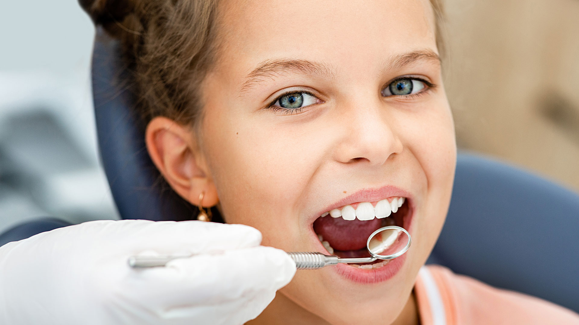 A young girl sitting in a dental chair, receiving dental care with a dental hygienist's hand visible holding a dental instrument near her mouth.