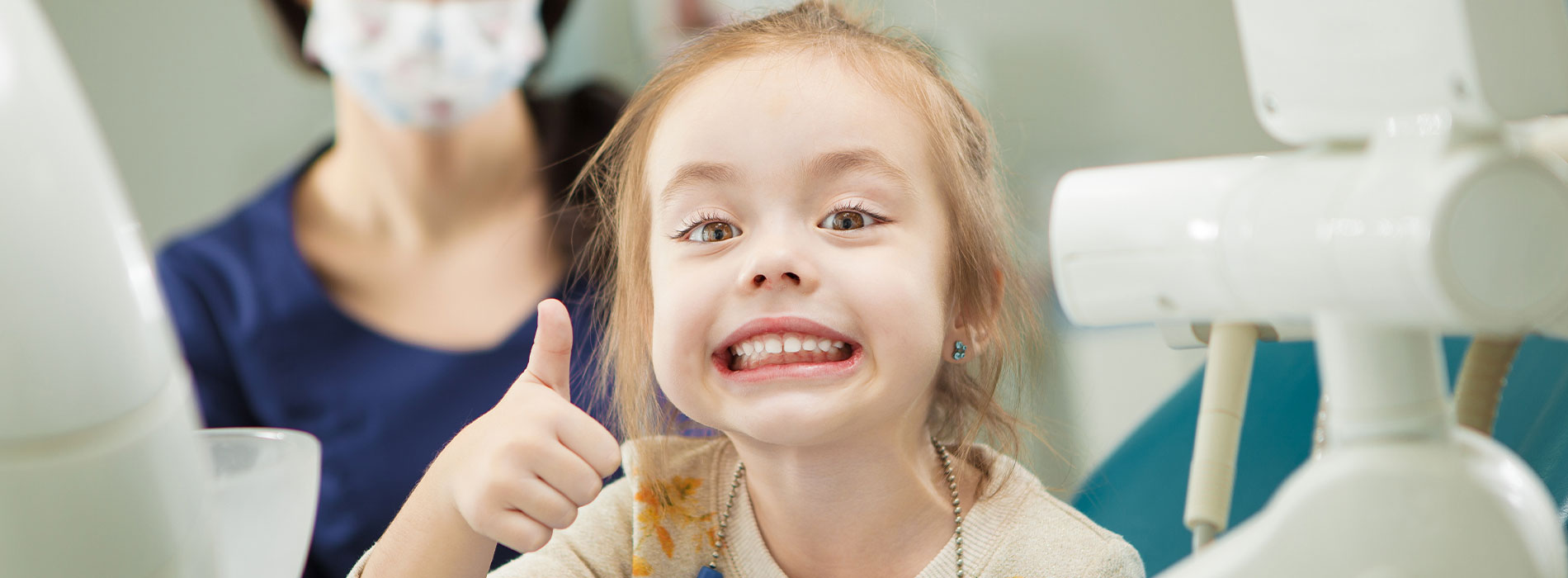 A young girl giving a thumbs-up sign while sitting in a dental chair with a dentist standing behind her, both in a dental office setting.
