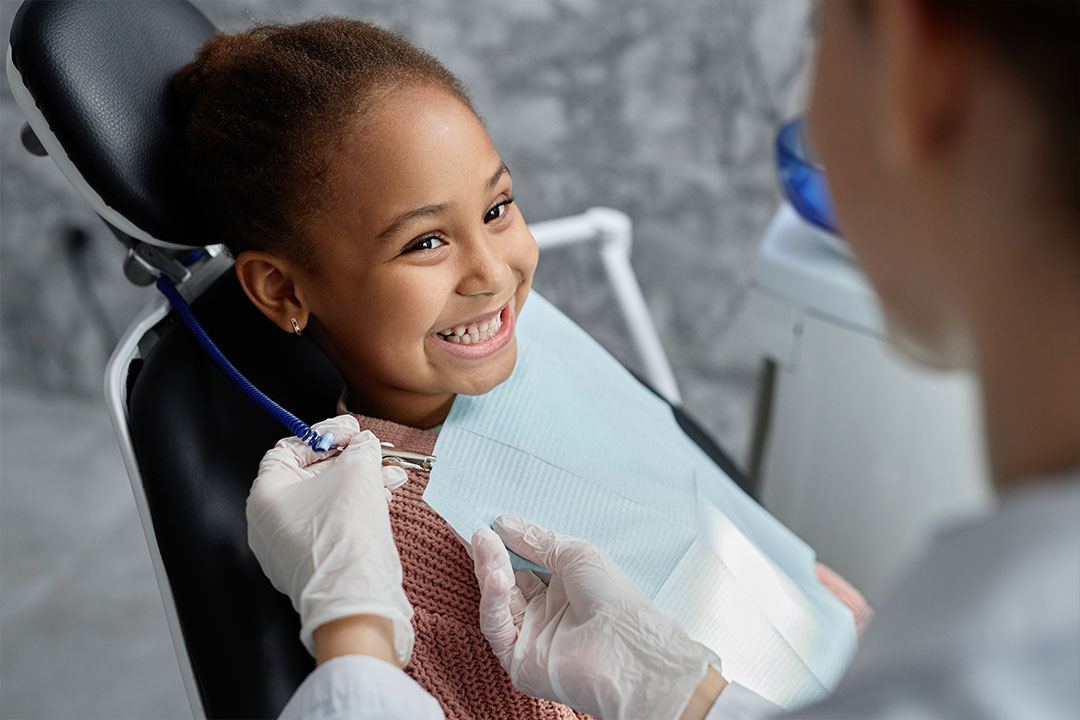 A young girl sitting in a dental chair with a smile, receiving dental care from a professional wearing gloves and a mask.