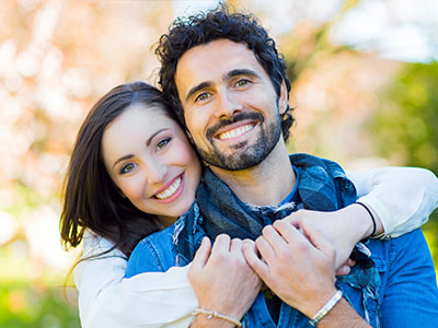 A man and woman are embracing each other with smiles, set against an outdoor background.