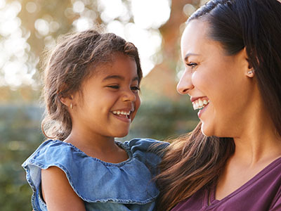 A woman and a young girl are smiling at each other outdoors during daylight.